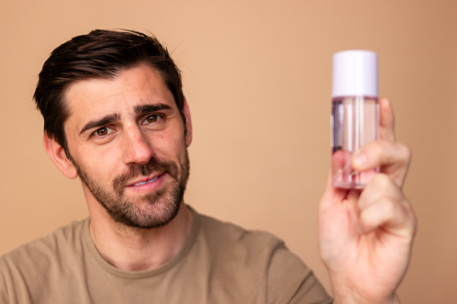 Male model presenting skincare cosmetic product bottle on beige background, focused on examining and holding up the grooming serum lotion for personal facial care routine and clear, healthy skin