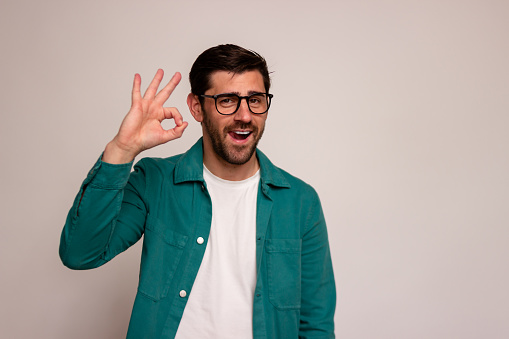 Smiling bearded man in glasses showing approval with an okay hand sign, isolated on a light backdrop
