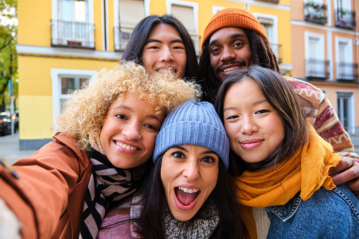 Multiracial friends group taking selfie pic with smartphone outside in autumn. Happy young people having fun walking on city street. Friendship concept with guys and girls enjoying outside.