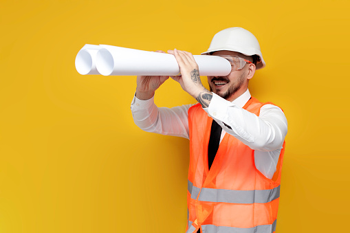 asian foreman in uniform looks into the distance on yellow isolated background, korean civil engineer in hard hat and goggles with papers and blueprints is watching forward