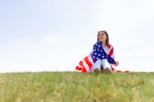 cute girl sitting on the grass and holding american flag in field on the lawn and looks into the distance, fourth of july concept