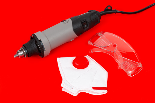 Corded mini drill, safety glasses and dust mask isolated on red background. Polishing and grinding protective gear concept.