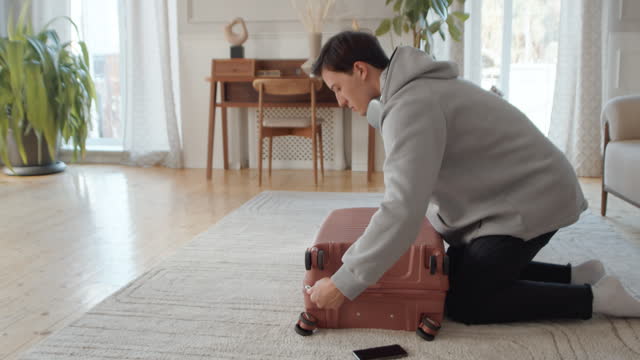 Young man neatly folding his clothes into a suitcase lying on the floor at home and zipping it during attentive preparation for the vacation
