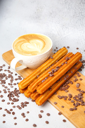 A cup of cappuccino and some churro sticks arranged neatly on a wooden cutting board.