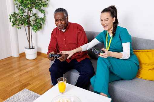 Laughter and learning echo through the room as a young female caregiver and a senior man delve into video games, exploring digital worlds together with joysticks in hand