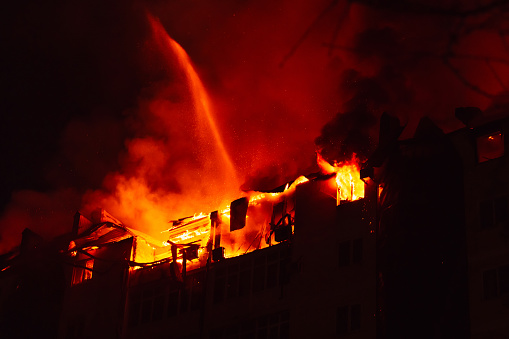 Burning house is engulfed in flames at night. Fire blazing in apartment building. Firefighters extinguish with water.
