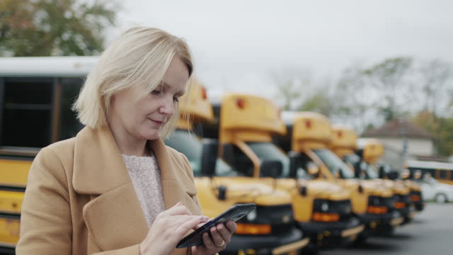 A teacher uses a smartphone, stands in the school yard against the background of yellow school buses. Side view