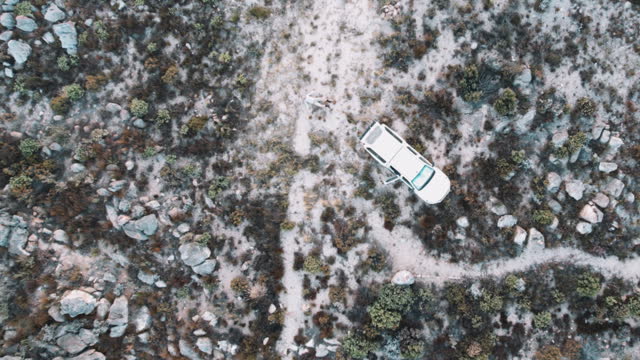 Directly above moving up drone shot man lying on his back making snow angels on the sand next to a vehicle parked in the Cederberg Wilderness