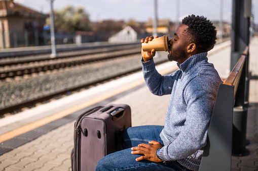 Happy man with suitcase enjoys drinking coffee while sitting on a bench at the railway station.