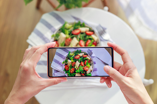 In the cropped image, an Asian woman can be seen using her smartphone to capture photos of a delicious homemade healthy salad before enjoying it at home. This represents the 'camera eats first' culture associated with both dining out and the trend of documenting food experiences on social media.