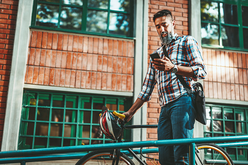 A handsome young man goes to the city with his bike, walking beside it and reading messages on smartphone.
