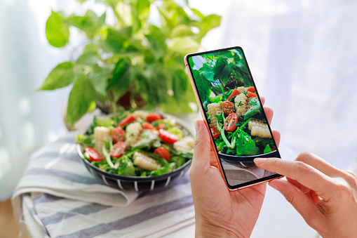 In the cropped image, an Asian woman can be seen using her smartphone to capture photos of a delicious homemade healthy salad before enjoying it at home. This represents the 'camera eats first' culture associated with both dining out and the trend of documenting food experiences on social media.