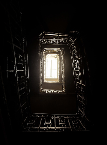 Looking up at the spiral staircase in old house. Dark staircase with metal bannister. Space filled with light coming from the upper floor.