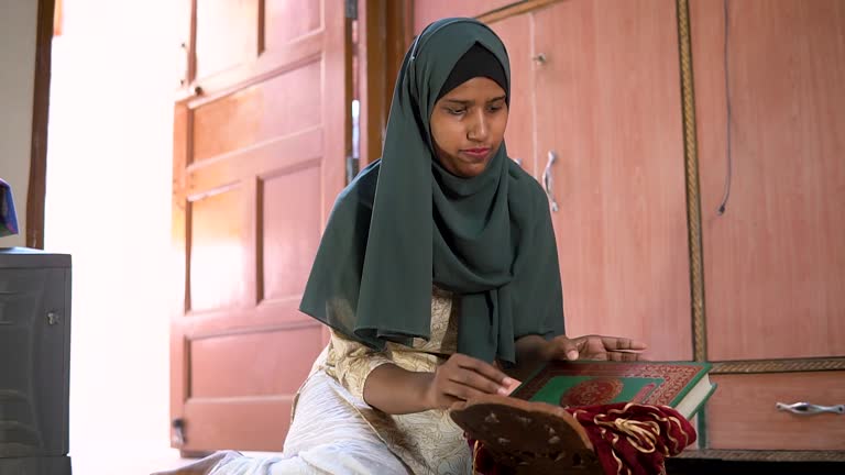 Indian Islamic woman in hijab sitting on prayer mat reading holy Quran in Ramadan.