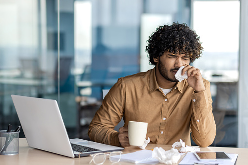 A sick young Indian man works in an office. Sitting at the table, not feeling well, holding a cup of drink and medicine, wiping his nose with a napkin from a runny nose.