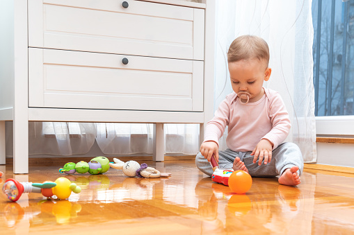 A baby with a pacifier in his mouth interacts with interest with toys while playing in the room.