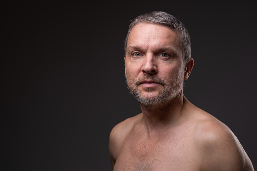 Slightly smiling face of a middle-aged man with a beard looking at the camera. Close-up portrait of a fifty-year-old man with a naked torso against a dark background.