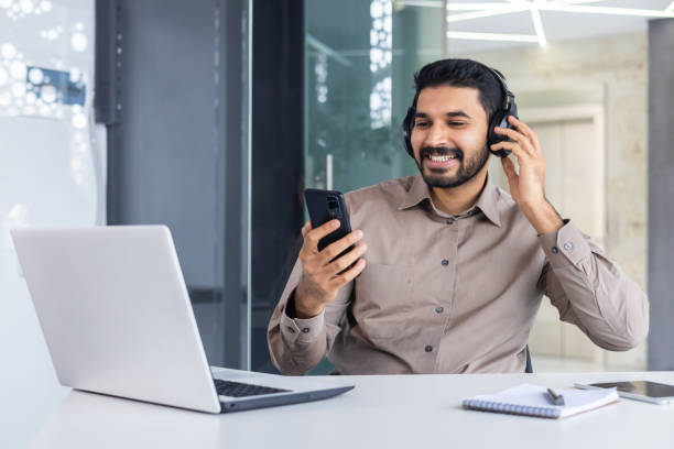 istock Satisfied successful hindu man at workplace inside office, man with phone in hands and headphones watching online video and listening to music audio podcasts, worker using laptop at work 2106777252