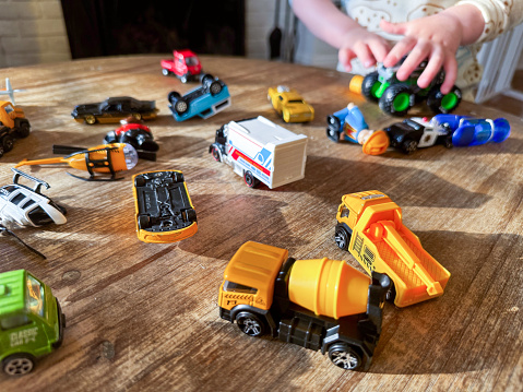 A child's hands play with toy construction vehicles and toy cars on a wooden table