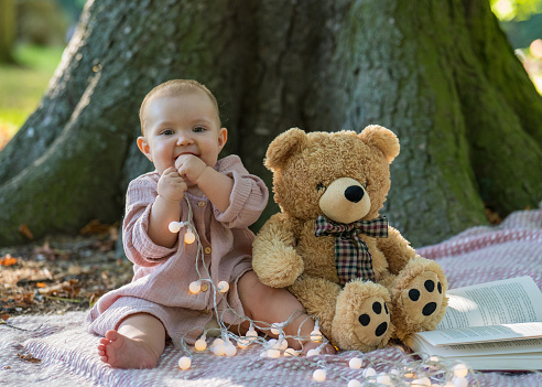Portrait of an adorable little girl in a beige dress sitting near a large tree and looking at the camera. He holds lanterns and chews them with pleasure. A large teddy bear sits next to the baby