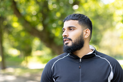 A thoughtful young adult male with a beard gazes into the distance in a serene park setting, surrounded by lush greenery on a bright sunny day.