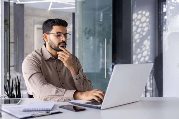 istock Man thinking inside office at workplace with laptop, businessman preparing financial report, typing on keyboard, financier accountant in glasses and shirt holding hand to chin 2106837343