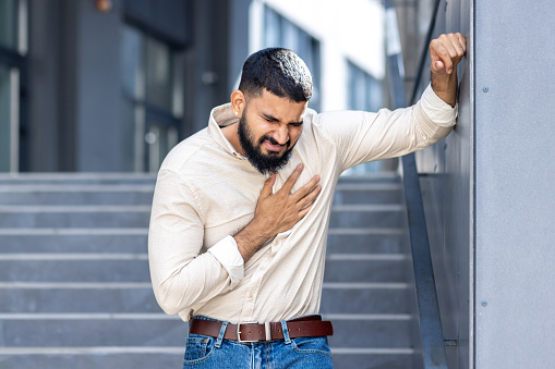 Panic and heart attack on the street. A young Indian man is standing near a building, leaning against a wall and holding his chest with his hand, feeling severe pain and needing medical help.