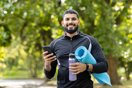 Happy, fit man holding a yoga mat and water bottle while using his smartphone in a sunny, green park.