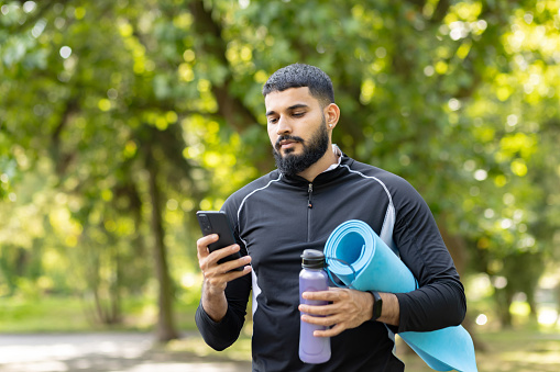 Focused male athlete with a yoga mat and water bottle using a smartphone in a sunlit park. Concept of technology in fitness and well-being.