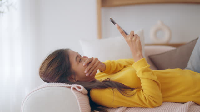 Happy young asian woman relax on comfortable couch at home texting messaging on smartphone, smiling girl use cellphone, browse wireless internet on gadget, shopping online from home