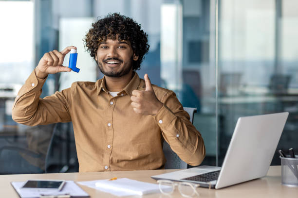Optimistic arab guy with curly hair holding inhaler and showing thumb up gesture while sitting by table with laptop. Allergic office manager using prescript medicine and feeling better during workday