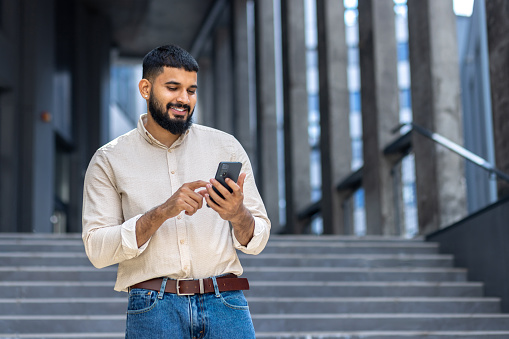 Joven estudiante indio de pie en el campus en las escaleras cerca del edificio con camisa y jeans y sonriendo usando el teléfono móvil photo