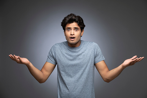 Young man spreading arms  as if not knowing what to do or surprised, isolated on gray background.