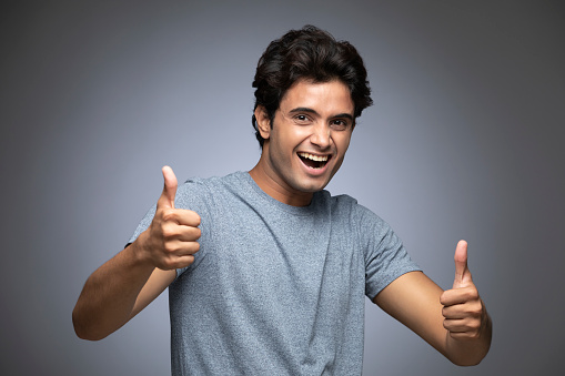 Young Indian man over isolated Grey background approving doing positive gesture with hand, thumbs up smiling and happy for success.