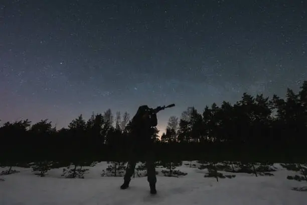 A soldier with a night vision and a rifle with a silencer and laser sight against the background of the starry sky in the forest in winter.High quality photo