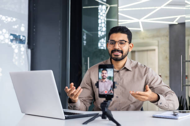 istock Successful business trainer teacher recording online training video course, man inside office at workplace using phone and tripod, sitting at desk with laptop 2106871924