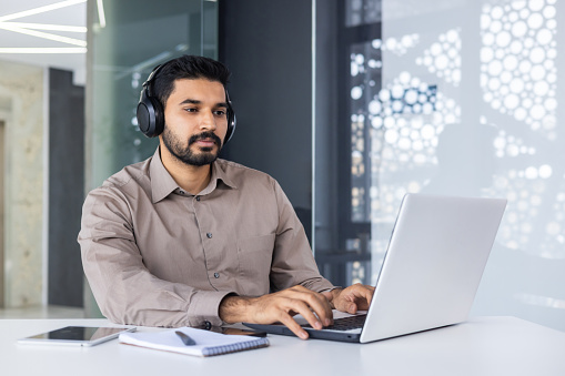 Focused latino male in formal shirt wearing modern headset and using portable computer while working by desk. Contemporary programmer getting ready for video meeting with professional garniture.