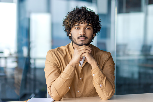 Serious hispanic startup leader supporting head with hands folded on desk and looking at camera on blurred background. Concerned entrepreneur thinking about profitable investment options and strategy.