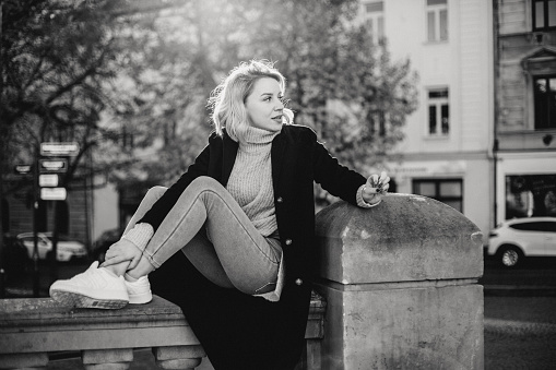 Stylish Woman in Black Coat and Jeans Sitting on Stone Wall. Casual Urban Fashion with White Shoes and Black Sweater. Relaxed Outdoor Portrait Perfect for Lifestyle and Fashion Concepts
