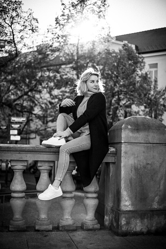 Chic Woman in Black Coat and White Shoes Sitting on Railing. Stylish Urban Fashion with Legs Crossed. Contemporary Casual Look Perfect for Lifestyle and Fashion Concepts. City Street Scene.