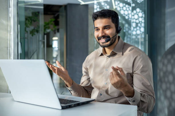 istock Young successful businessman at workplace talking remotely sitting at table inside office, man with headset using laptop for video call, online customer support worker 2106887386