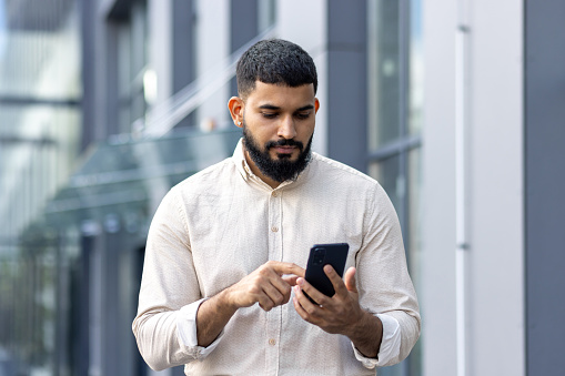 Photo of a serious Indian young man walking down the street near office buildings and using a mobile phone.