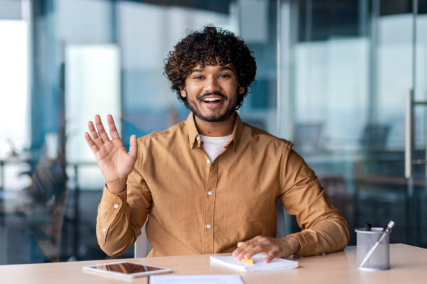 istock Smiling hispanic employee making hello gesture while keeping hand on table with daybook, pen and tablet. Pleasant bearded man welcoming colleague in meeting room before beginning of discussion 2106926672