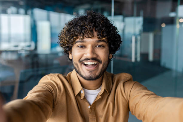 istock Smiling indian office worker looking at camera talking with toothy smile on background of glassy walls. Happy smm manager creating content and recording video for new post in company social media 2106939524