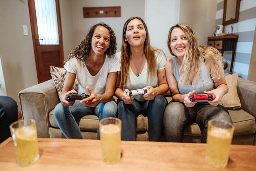 Three joyful women sitting on a couch, enjoying video games together, with refreshing drinks on the table in a cozy living room.