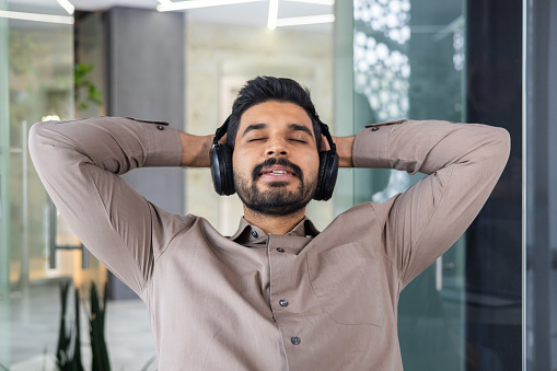 Portrait of relaxed indian man in headphones leaning on back of chair with closed eyes on blurred background. Calm manager resting in office with hands behind head and listening to soothing playlist.