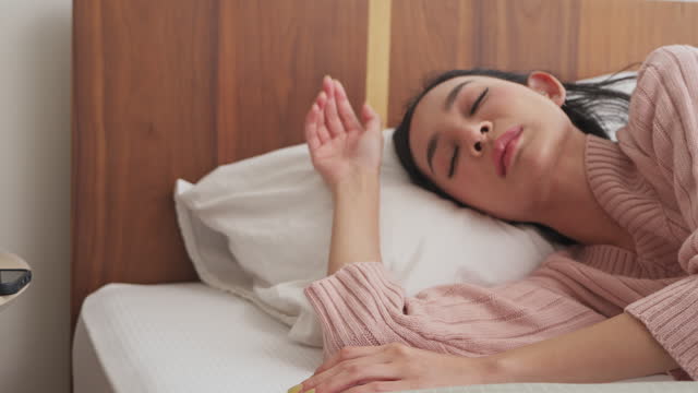 Young Asian woman in a soft pink sweater lying in bed with a headache, hand on forehead, with a serene expression
