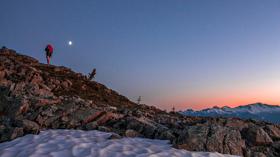 Backpacker hikes mountain ridge at dawn, moon in distance