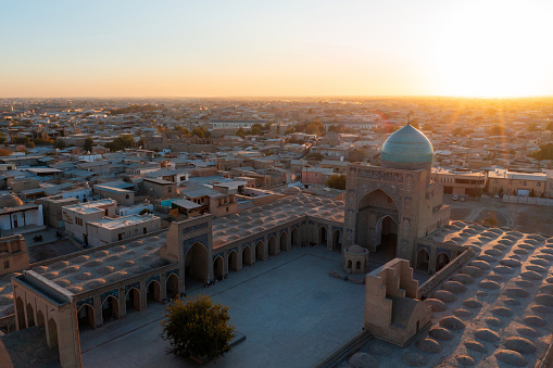 Bukhara Sunset over the famous Kalyan Poi Kalon, Poi Kalan or Po-i-Kalyan and Kalon Mosque with courtyard in the old town of Bukhara - Buxoro - Бухорo. Aerial Drone Point View against the Sun at Sunset. Itchan Kala, Bukhara, Khorezm Region, Uzbekistan, Central Asia.