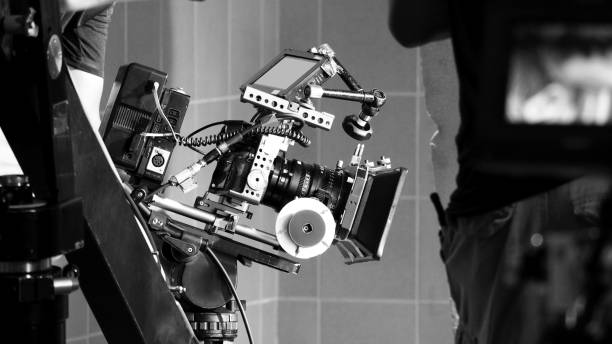 istock Back angle view of high definition digital video camera which recording or shooting tv commercial production on pan tilt shift professional head tripod at outdoor location and film crew team with set. 2107262395